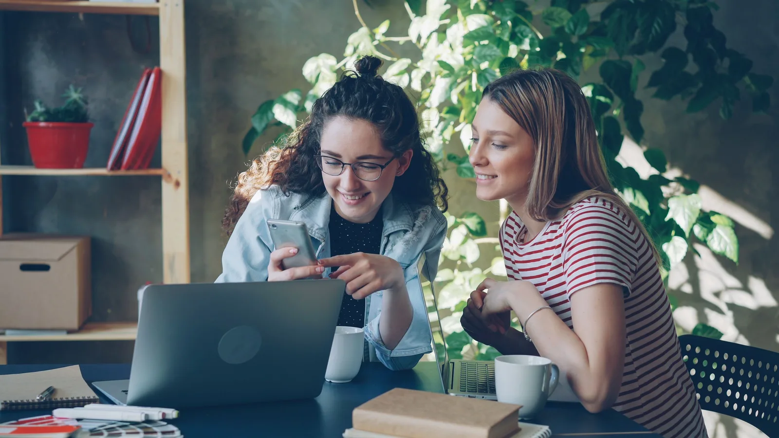 Two women using a laptop and smartphone in a cozy, plant-filled office setting. 9 social media strategy checks before signing off